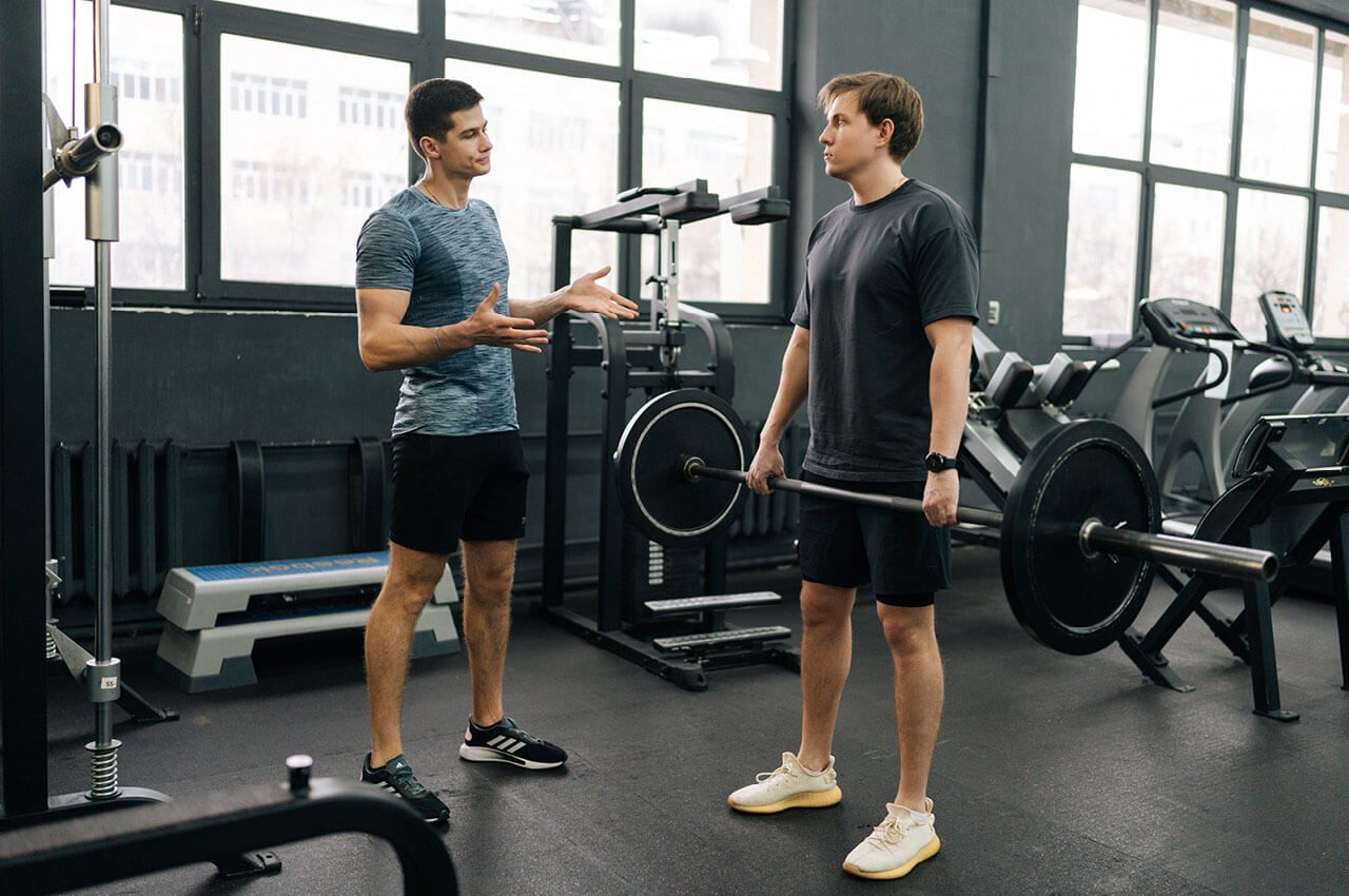 Trainer erklärt einem Mann die richtige Übungsausführung beim Heben einer Langhantel in einem Fitnessstudio.