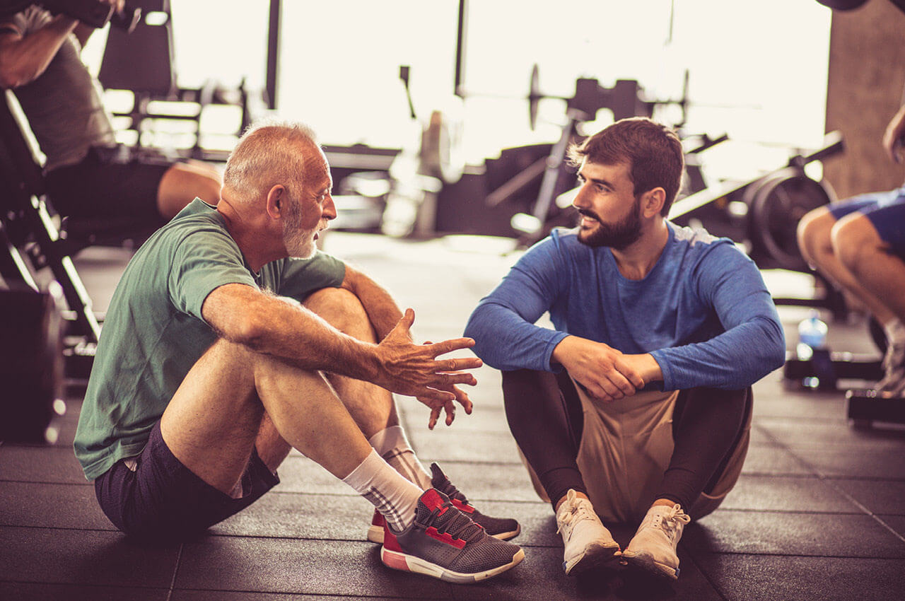 Ein alter und ein junger Mann sitzen in einem Fitnessstudio auf dem Boden und unterhalten sich