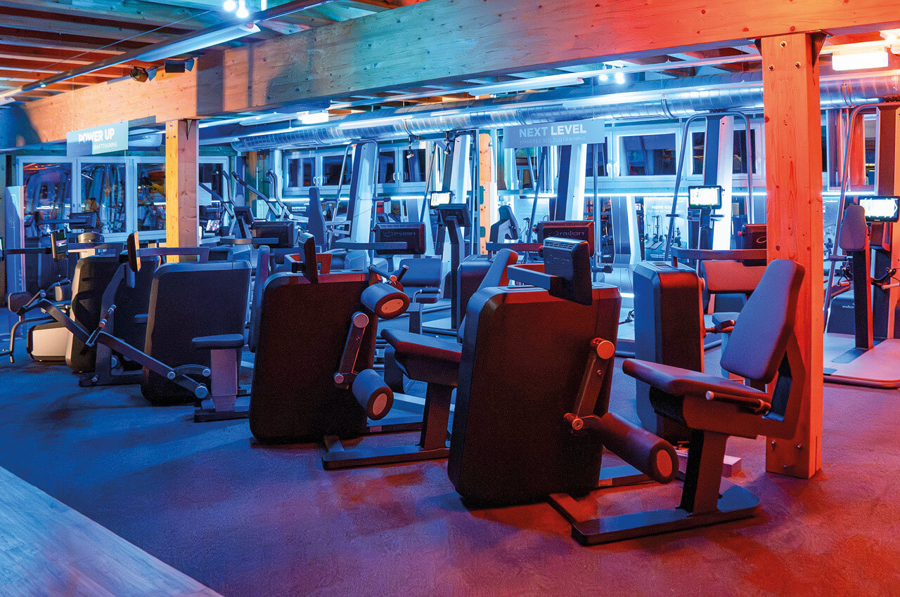Fitnessstudio &bdquo;Gesund und Fit&ldquo; mit vielen verschiedenen Ger&auml;ten, rotem und blauem Licht und Holzbalken im Raum