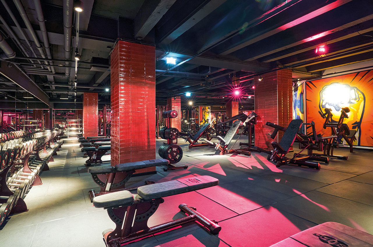 Fitnessstudio mit roten S&auml;ulen, Hantelb&auml;nken und Kraftger&auml;ten, beleuchtet durch farbiges Neonlicht; an der Wand ein gro&szlig;fl&auml;chiges Graffiti.