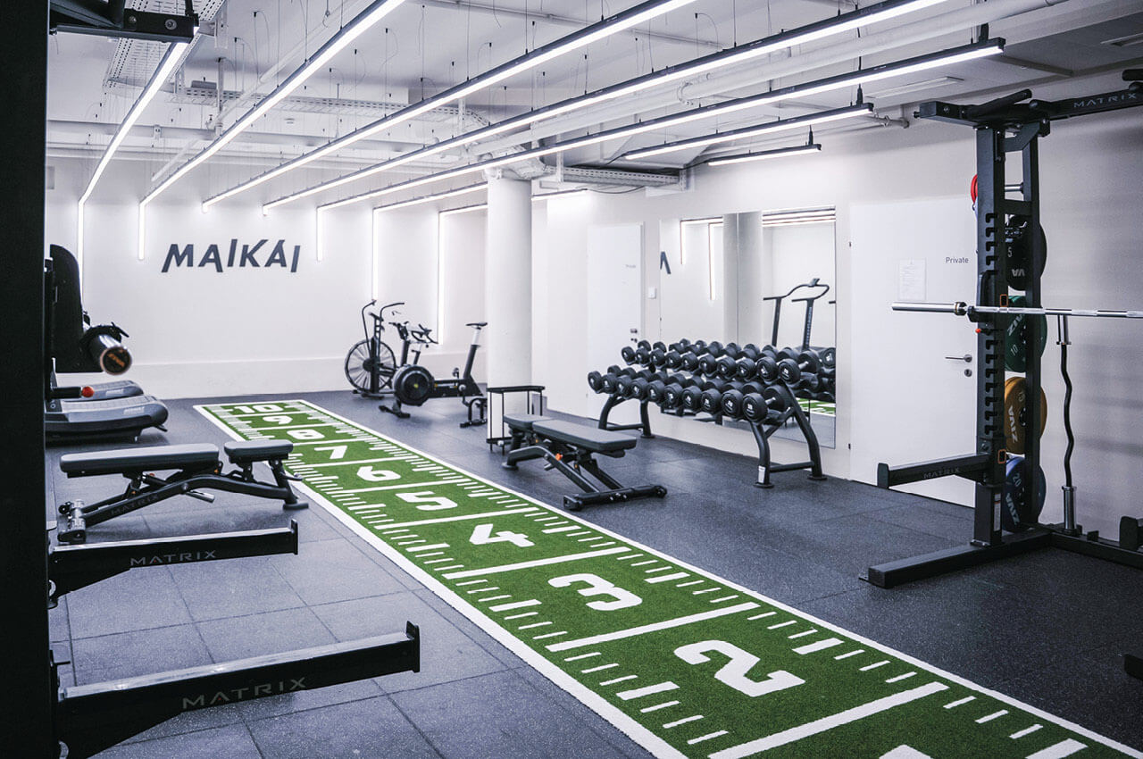 Helles Fitnessstudio mit Hanteln, Trainingsger&auml;ten und gr&uuml;nem Kunstrasen mit aufgedruckten Markierungen f&uuml;r Functional Training, an der Wand der Schriftzug Maikai.
