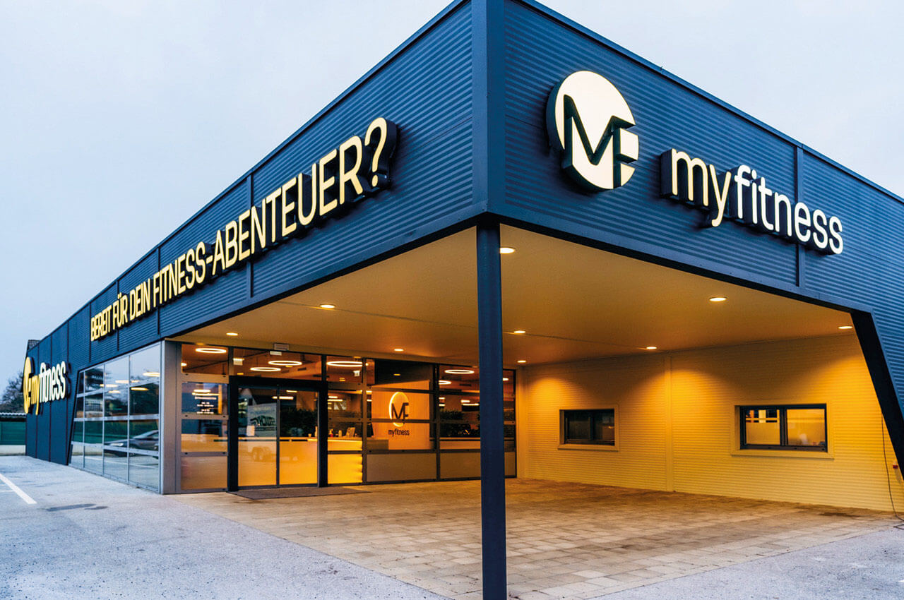 Au&szlig;enansicht eines modernen Fitnessstudios mit der Leuchtschrift &sbquo;myfitness&lsquo; und dem Slogan &sbquo;Bereit f&uuml;r dein Fitness-Abenteuer?&lsquo; &uuml;ber dem Eingang.
