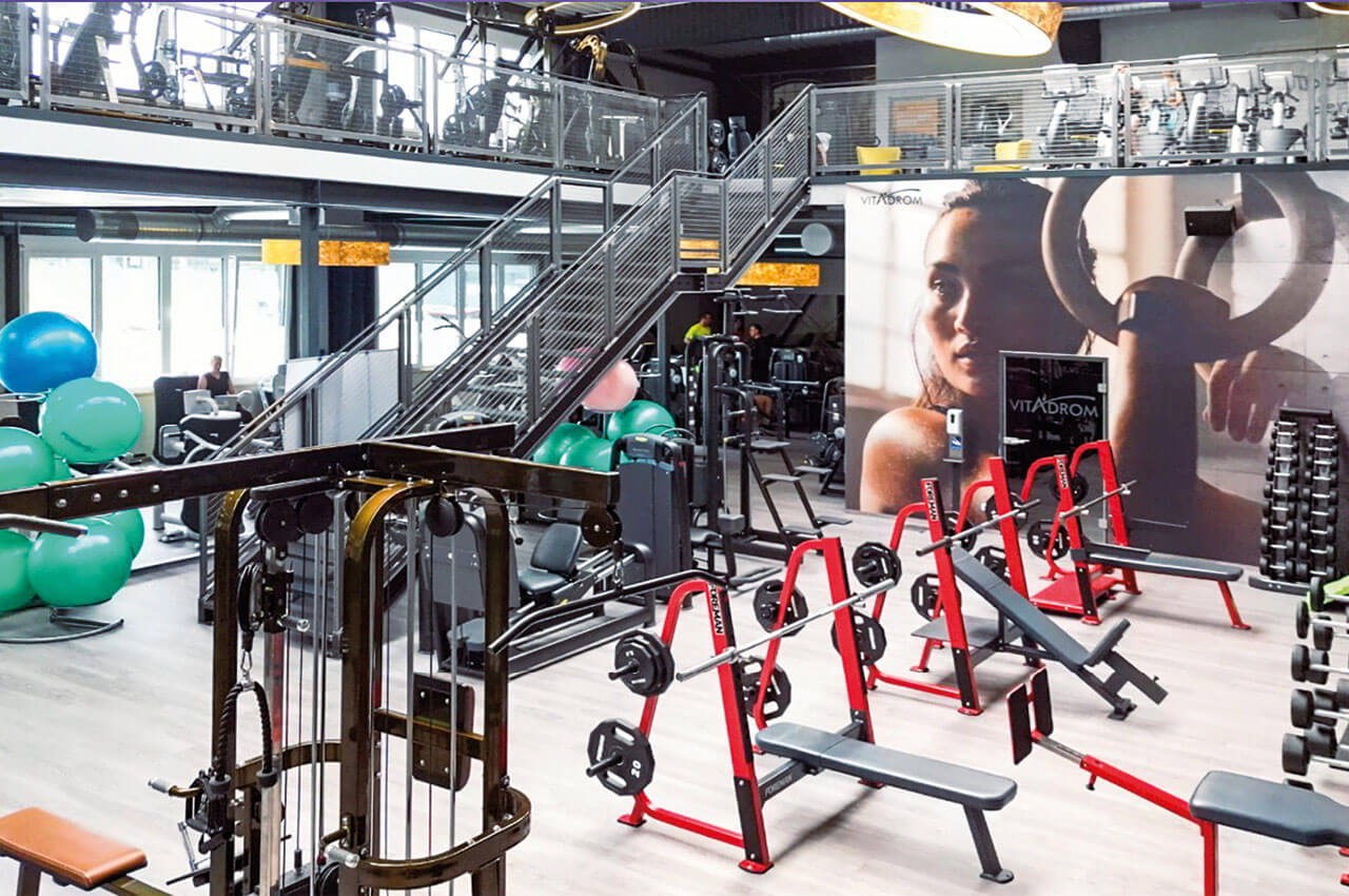 Gro&szlig;z&uuml;giges Fitnessstudio mit Freihantelbereich, roten Hantelstationen, Fitnessger&auml;ten und einem Wandbild mit einer Sportlerin, dar&uuml;ber ein Trainingsbereich auf der Galerie.