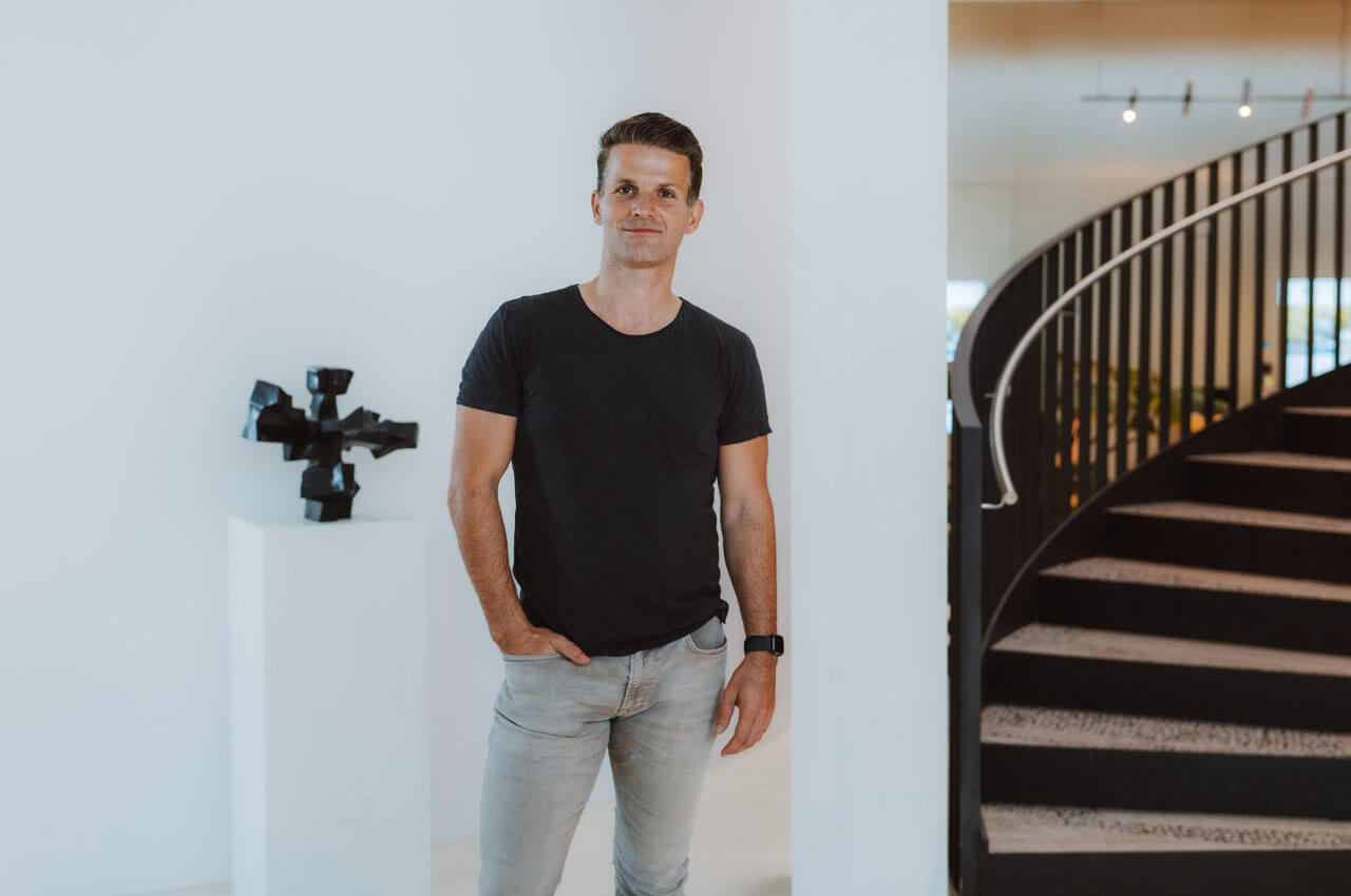 Ein Mann in einem schwarzen T-Shirt und hellen Jeans steht l&auml;ssig mit einer Hand in der Hosentasche in einem modernen, hellen Geb&auml;ude. Im Hintergrund ist eine geschwungene, dunkle Treppe sowie eine abstrakte schwarze Skulptur auf einer wei&szlig;en S&auml;ule zu sehen.