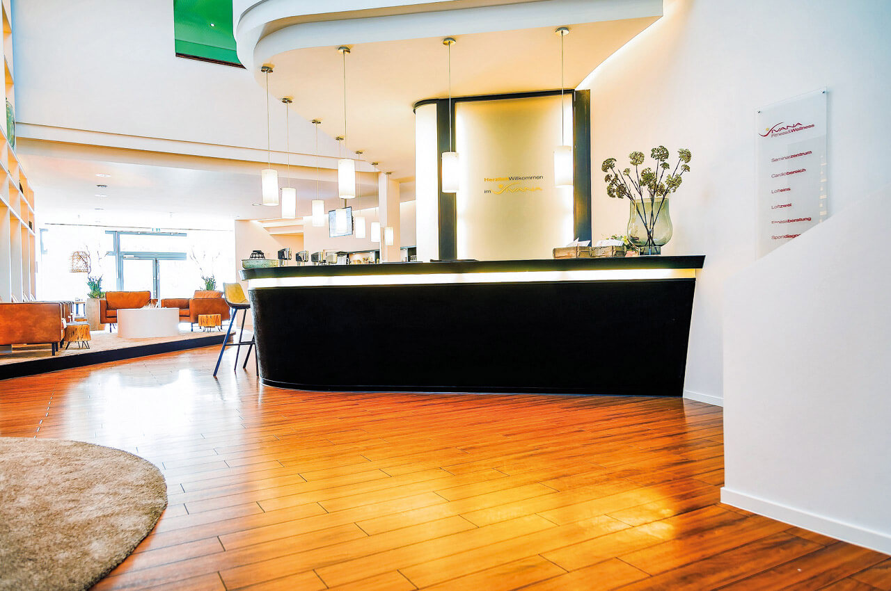 Service, der sich nach Urlaub anfühlt Ein heller und moderner Empfangsbereich eines Fitness- und Wellnessstudios mit hochwertigem Holzfußboden. Im Zentrum steht ein geschwungener, schwarzer Tresen mit indirekter Beleuchtung, über dem mehrere schlichte Pendelleuchten hängen. Im Hintergrund ist ein gemütlicher Loungebereich mit Ledersesseln erkennbar, während an der rechten Wand eine Infotafel mit den verschiedenen Studiobereichen angebracht ist.