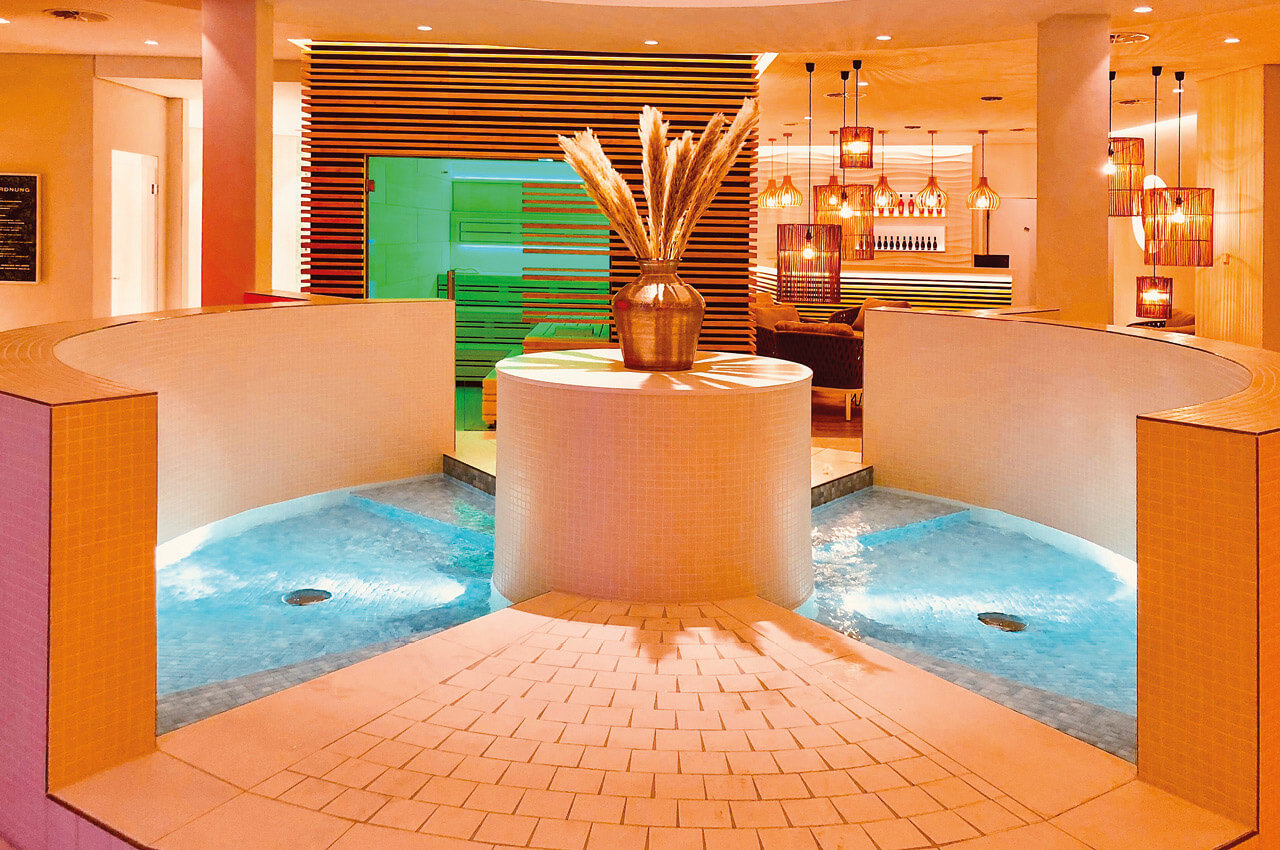 Service, der sich nach Urlaub anfühlt - 2 Ein stimmungsvoll beleuchteter Wellnessbereich mit einem runden Becken im Vordergrund, das mit hellblauem Wasser gefüllt ist. In der Mitte des Beckens steht eine dekorative goldene Vase mit hohen Gräsern. Im Hintergrund ist eine Holzsauna mit grünlicher Innenbeleuchtung sowie ein Barbereich mit hängenden Korbgeflecht-Leuchten zu erkennen.