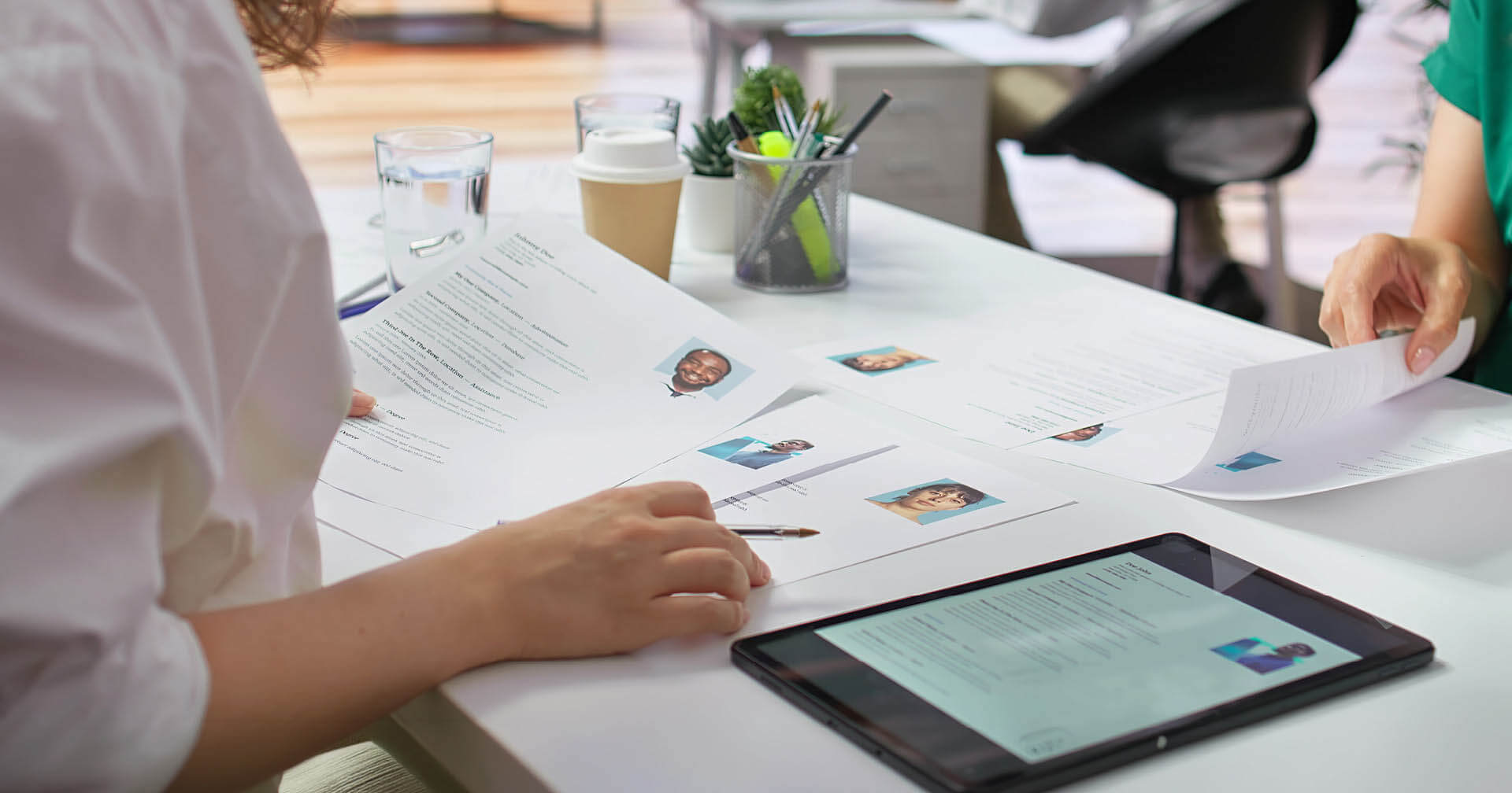 Blick über die Schulter einer Person am Schreibtisch während eines Bewerbungsgesprächs oder einer Personalauswahl. Auf dem hellen Tisch liegen ausgedruckte Lebensläufe mit Passfotos sowie ein Stift, Gläser, ein Kaffeebecher und ein Tablet, das ebenfalls Unterlagen zeigt.