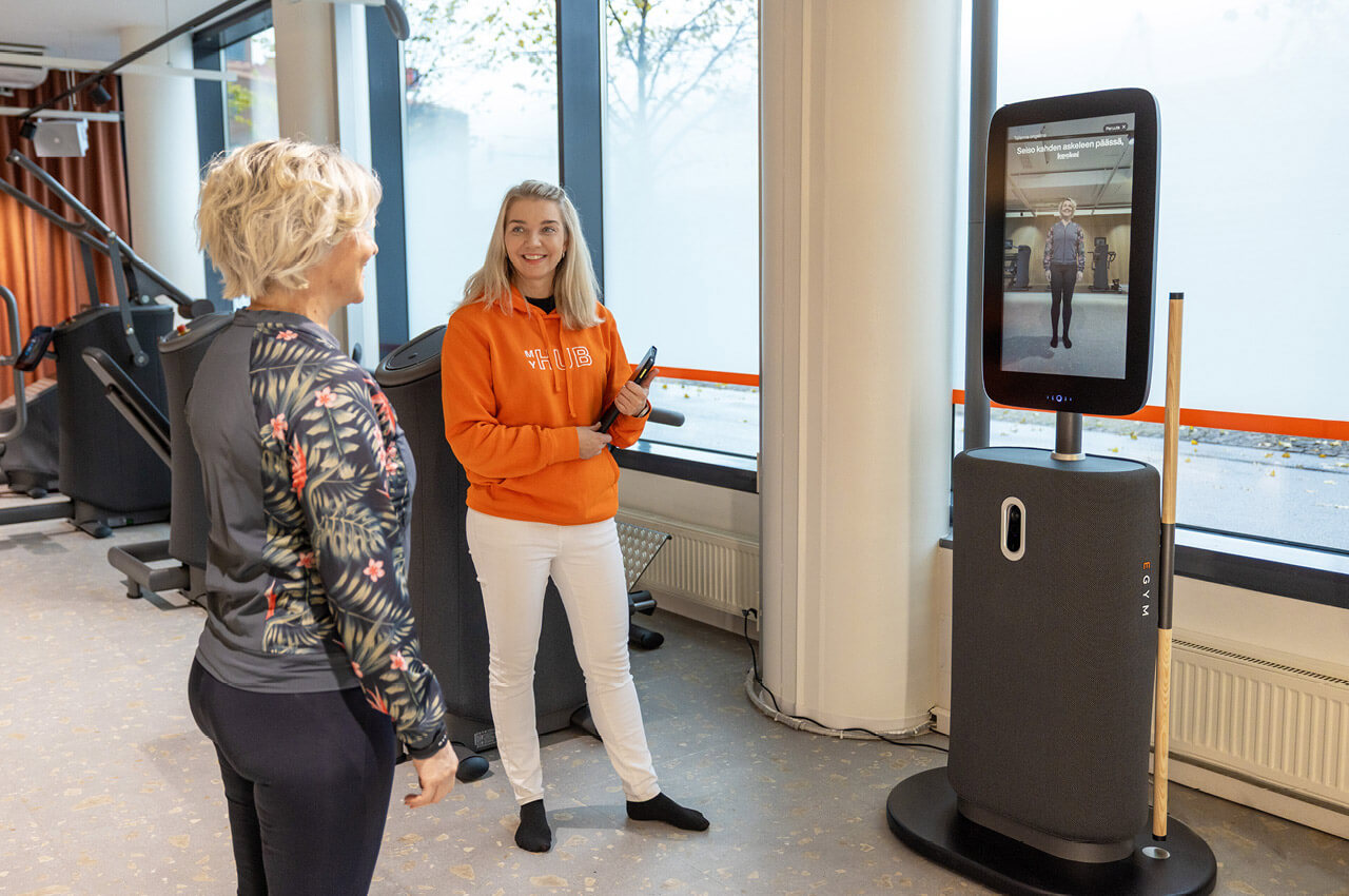 Eine Trainerin in einem orangefarbenen Kapuzenpullover mit der Aufschrift &bdquo;MyHub&ldquo; steht neben einer Frau vor einem EGYM Fitness Hub (K&ouml;rperscanner). Auf dem gro&szlig;en vertikalen Bildschirm des Hubs ist die Silhouette der Frau f&uuml;r die Haltungsanalyse zu sehen.