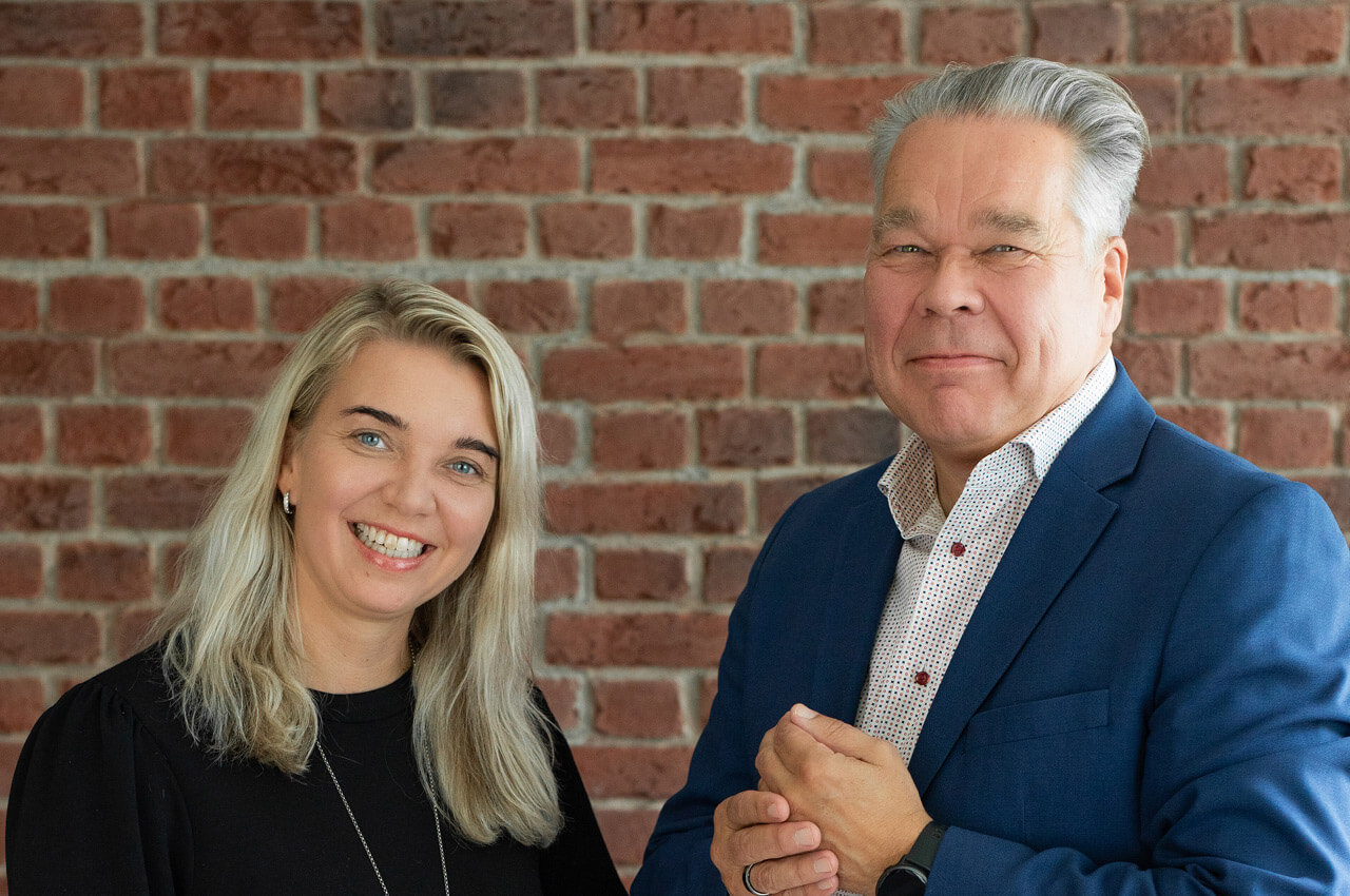 Portr&auml;tfoto einer l&auml;chelnden Frau mit blonden Haaren und eines &auml;lteren Mannes im blauen Sakko vor einer rustikalen roten Backsteinwand.