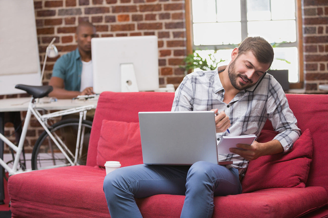 Ein Büromitarbeiter sitzt auf der Couch mit einem Laptop und einem Smartphone am Ohr.