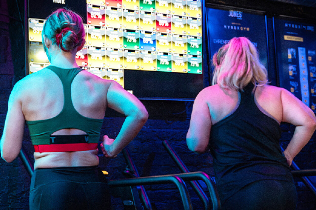 Zwei Frauen trainieren auf Laufbändern in einem Fitnessstudio und blicken auf große Bildschirme, die bunte Trainings- und Leistungsdaten anzeigen.