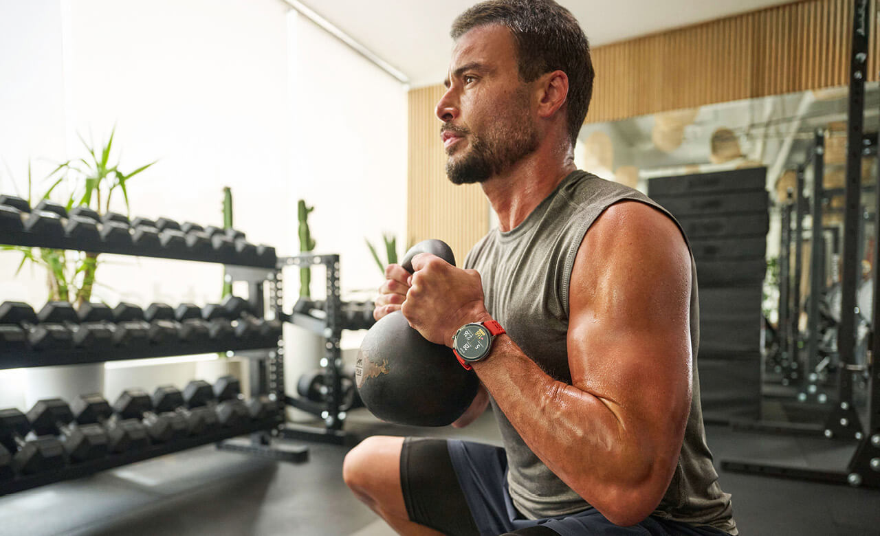 Mann im Fitnessstudio trainiert mit einer Kettlebell in der Hocke und trägt eine Smartwatch am Handgelenk, im Hintergrund sind Hanteln und Fitnessgeräte zu sehen.