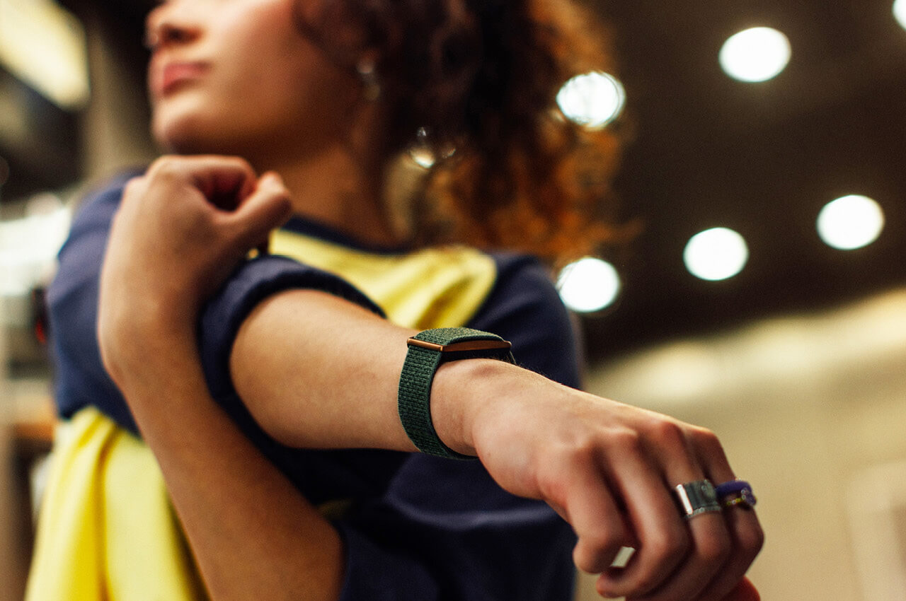 Nahaufnahme einer Person während einer Dehnübung, wobei der Arm mit einem dunkelgrünen, textilen Fitness-Tracker-Armband sichtbar ist. Die Person trägt ein dunkelblaues Oberteil über einem gelben Shirt und hat lockiges, rötliches Haar. Im unscharfen Hintergrund sind helle, kreisförmige Lichter zu sehen.