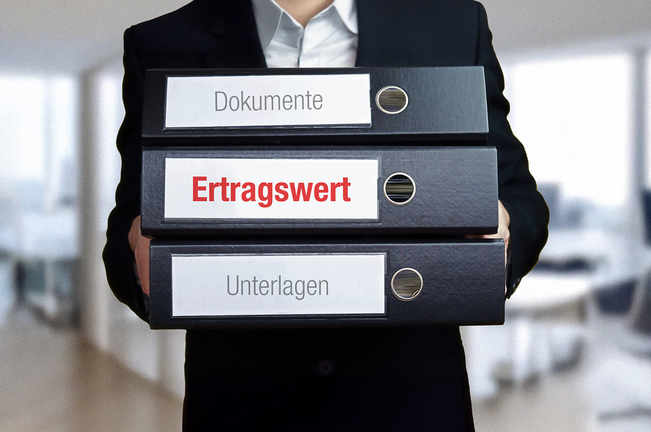 Ein in dunklem Anzug gekleideter Mann hält einen Stapel von drei schwarzen Aktenordnern vor der Brust. Die Beschriftungen der Ordner von oben nach unten lauten: "Dokumente", in roter und großer Schrift "Ertragswert" und "Unterlagen". Der Hintergrund zeigt ein leicht unscharfes, modernes Büro.