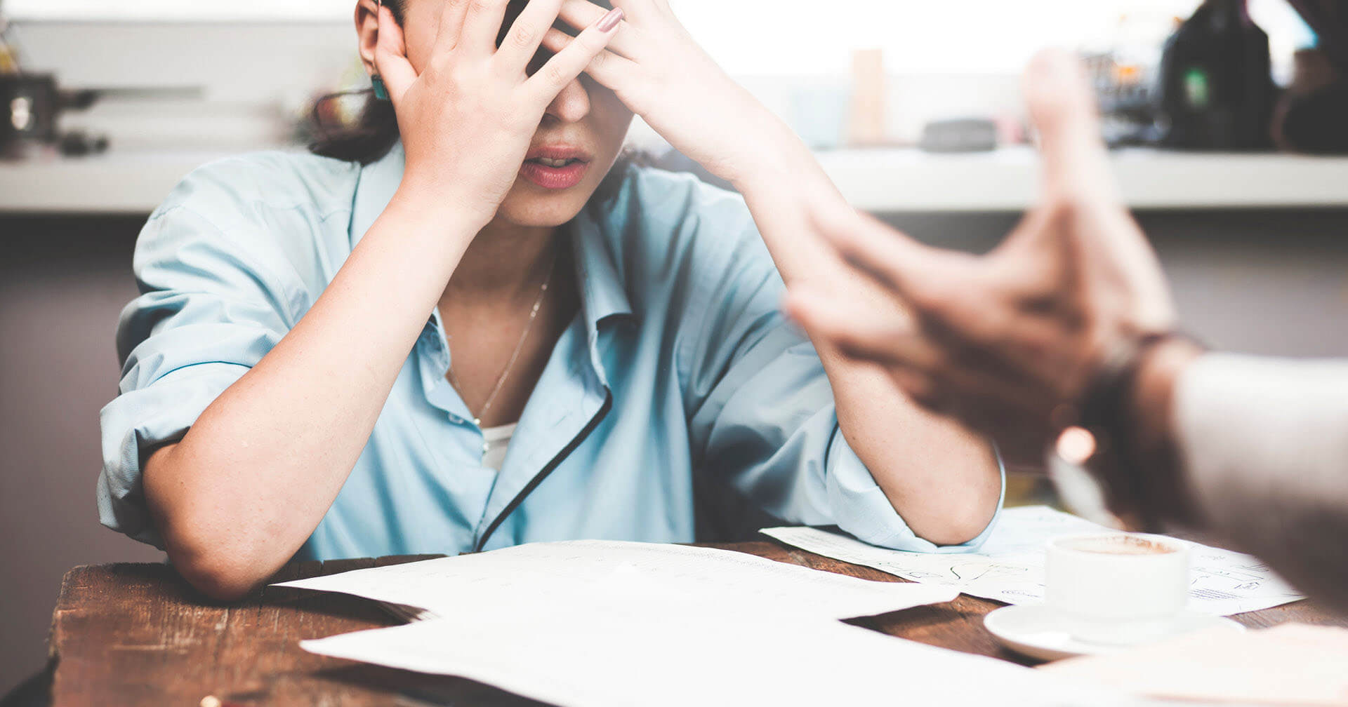 Nahaufnahme einer Person am Schreibtisch, die verzweifelt beide Hände vors Gesicht schlägt. Sie trägt ein hellblaues Hemd. Im unscharfen Vordergrund gestikuliert eine andere Hand fordernd. Dokumente und eine Kaffeetasse liegen auf dem Holztisch.