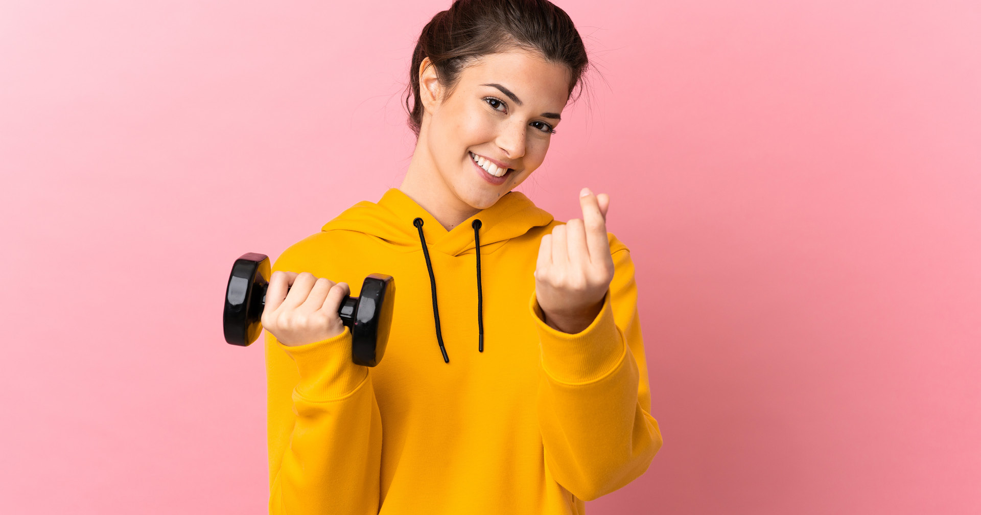 Fitnessstudiomitglied steht mit Hantel in der Hand vor einem rosa Hintergrund. 