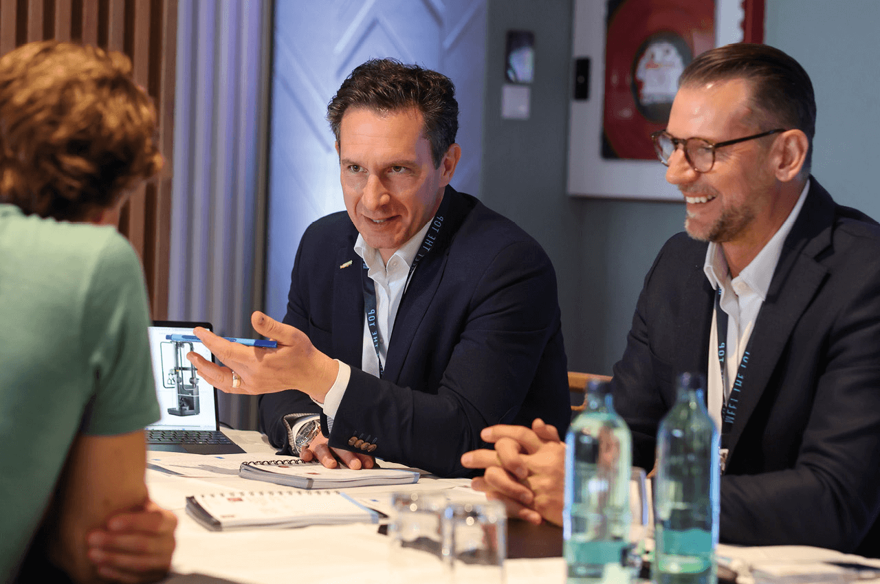 An einem Meetingtisch sitzen 3 Herren und verhandeln.