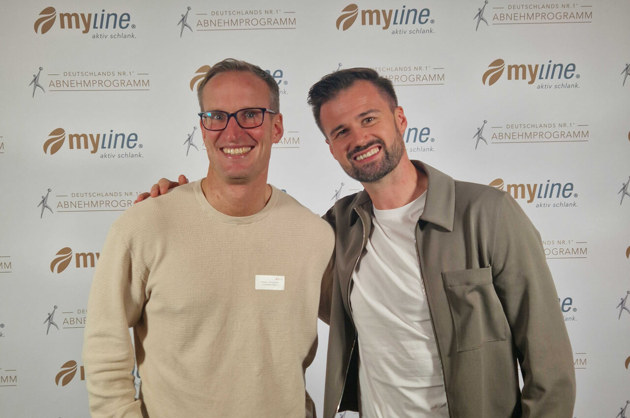 Zwei lächelnde Männer stehen vor einer myline-Sponsorenwand mit dem Slogan "DEUTSCHLANDS NR. 1® ABNEHMPROGRAMM". Links steht Conny Wilser (BODYMEDIA) in einem beigefarbenen Pullover und Brille. Rechts posiert Eric Herrforth (myline), der eine khakifarbene Jacke trägt.