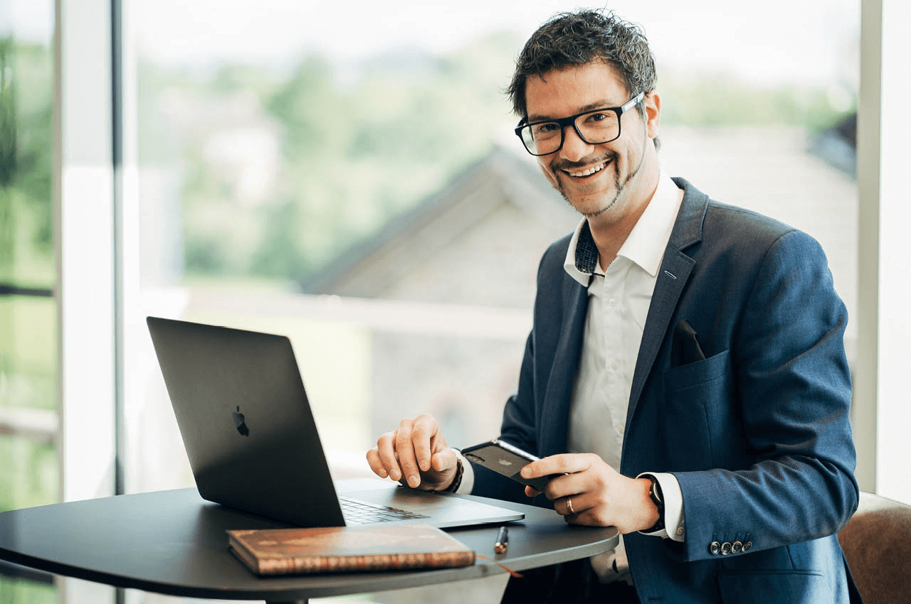 Ein Physiotherapeut sitzt an einem Schreibtisch mit einem Laptop und lächelt in die Kamera. 