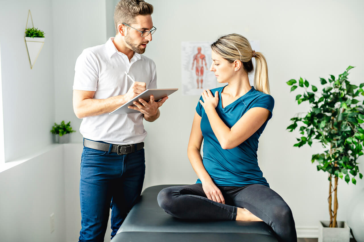 Ein Physiotherapeut mit Brille und wei&szlig;em Poloshirt macht sich mit einem Eingabestift Notizen auf einem Tablet. Vor ihm sitzt eine Patientin in Sportkleidung auf einer Behandlungsliege und deutet auf ihre schmerzende Schulter. Die Szene findet in einem hellen Behandlungsraum mit Zimmerpflanzen statt.