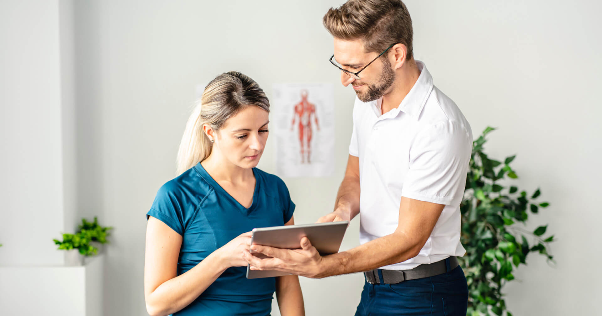 Ein Mann in weißem Poloshirt und eine Frau in blauem Sportshirt stehen nebeneinander in einer Praxis und blicken gemeinsam auf ein Tablet. Der Mann zeigt auf das Display, während im Hintergrund eine Pflanze und ein anatomisches Poster zu sehen sind.