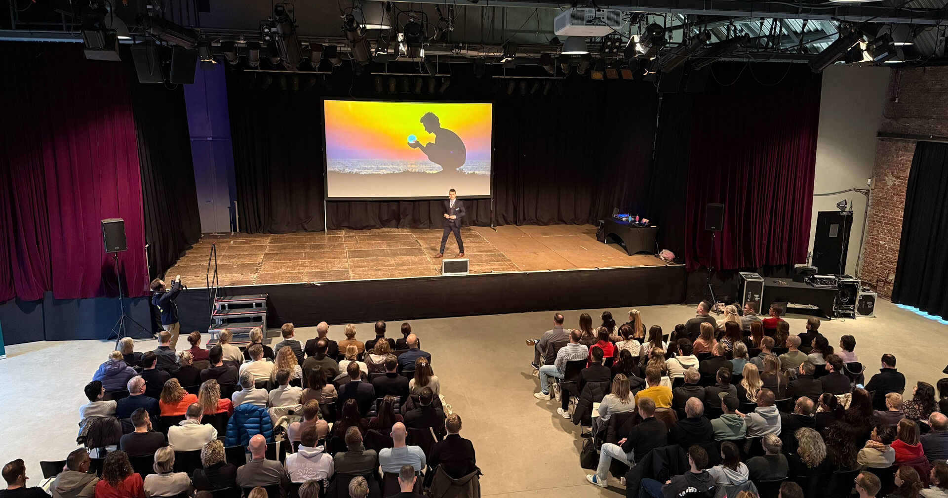 Weitwinkelaufnahme eines gut besuchten Vortragssaals. Ein Redner steht auf einer Bühne vor einer großen Leinwand, die eine Silhouette vor einem Sonnenuntergang zeigt. Das Publikum sitzt in Reihen und blickt zur Bühne; die Atmosphäre ist professionell und durch Bühnenscheinwerfer beleuchtet.