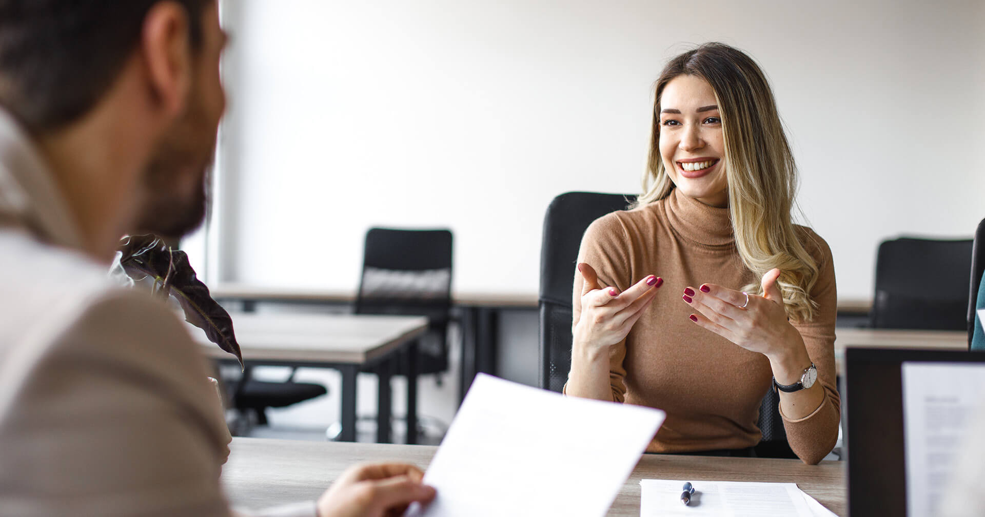 Eine lächelnde junge Frau mit blonden Haaren im braunen Rollkragenpullover sitzt in einem modernen Büro an einem Tisch und gestikuliert lebhaft während eines Gesprächs. Ihr Gegenüber ist nur von hinten im Anschnitt zu sehen und hält ein Dokument in der Hand.