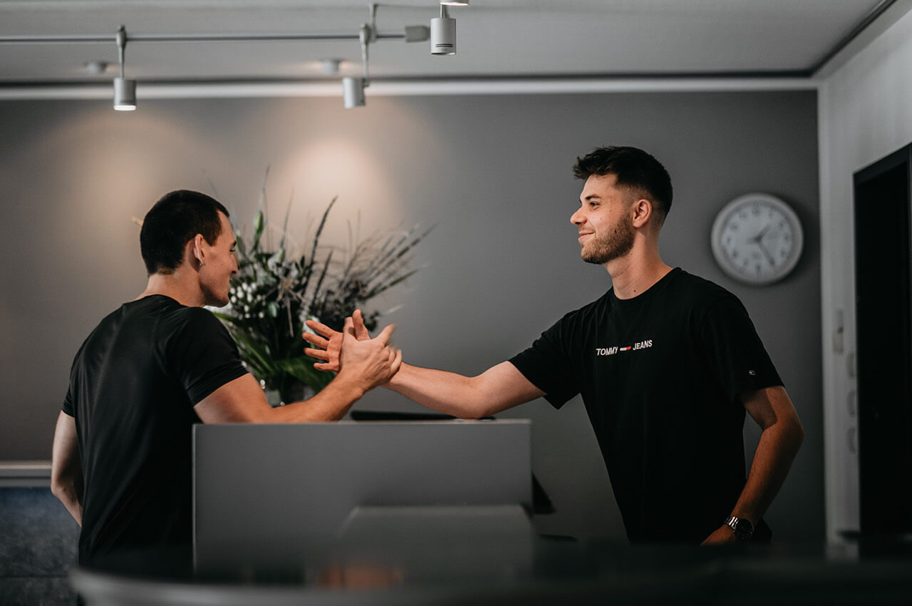 Zwei junge M&auml;nner in schwarzen T-Shirts begr&uuml;&szlig;en sich herzlich mit einem Handschlag &uuml;ber den Tresen eines Empfangsbereichs hinweg.