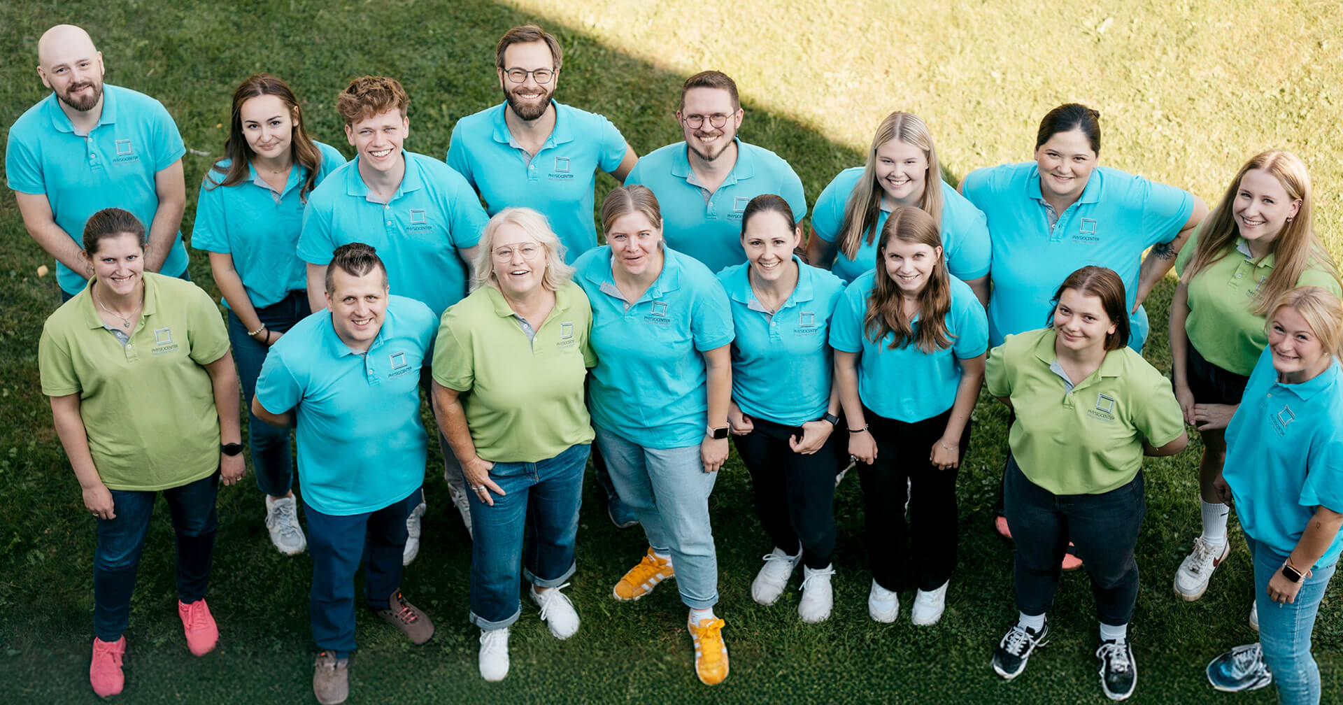 Eine Draufsicht auf ein großes, gemischtes Team von 15 Mitarbeitern einer Praxis, die in blauen und lindgrünen Poloshirts auf einer Rasenfläche für ein Gruppenfoto posieren. Die Gruppe steht eng beieinander und lächelt in die Kamera.