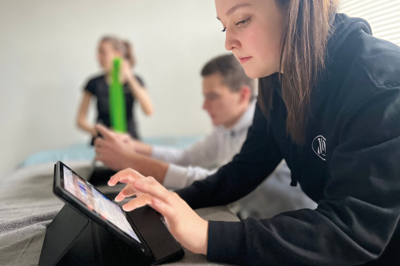 Eine Physiotherapeutin blickt, gebückt auf ein Tablet im Hintergrund sind verschwommen zu sehen zwei weitere Physiotherapeuten.