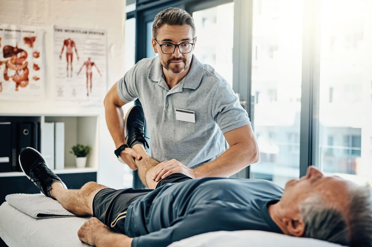 Physiotherapeut f&uuml;hrt eine Mobilisations&uuml;bung am Bein eines auf der Behandlungsliege liegenden Patienten durch.