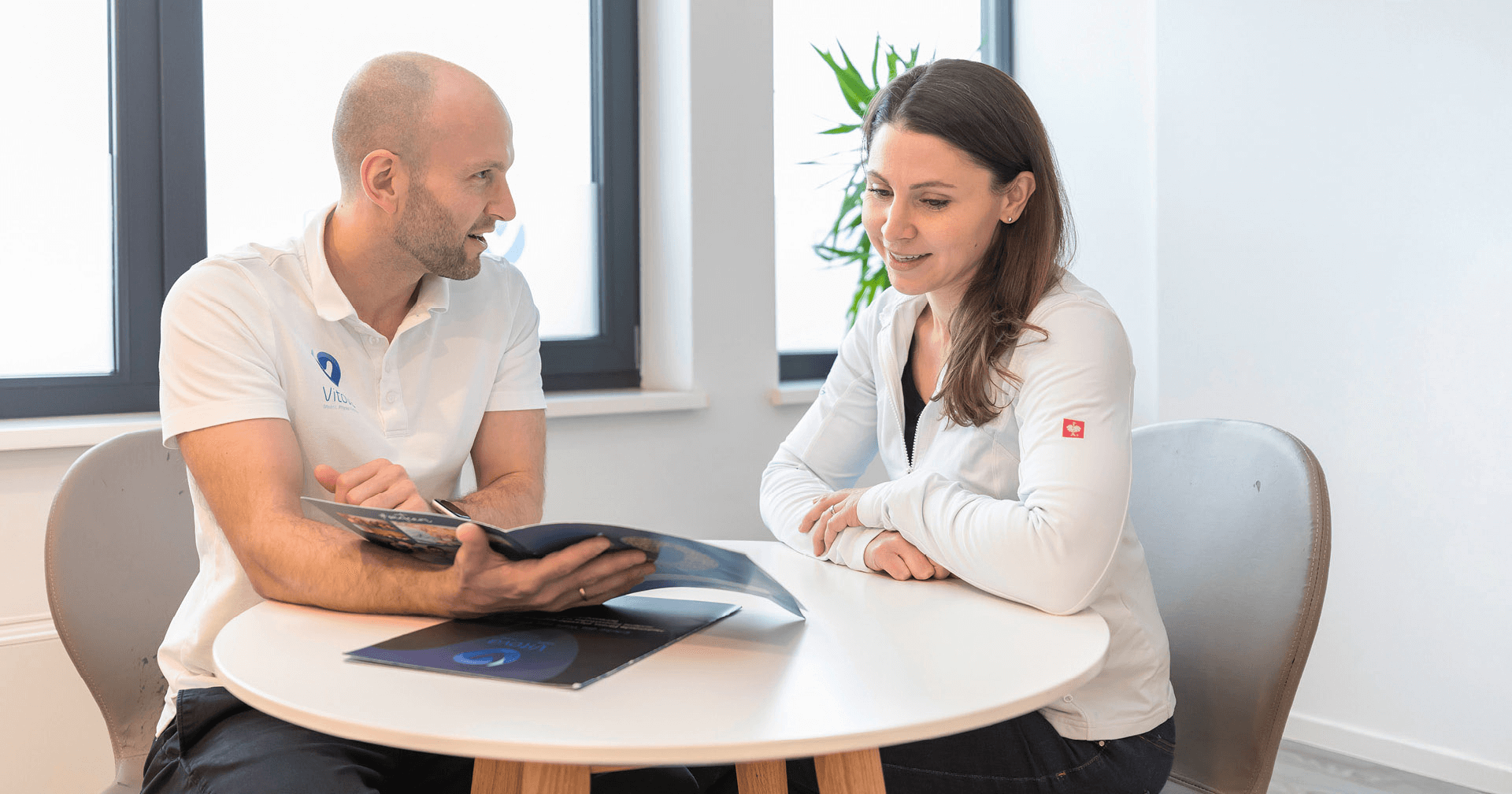 Ein Physiotherapeut sitzt am Tisch mit einer Mitarbeiterin und erklärt ihr anhand eine Broschüre etwas. 
