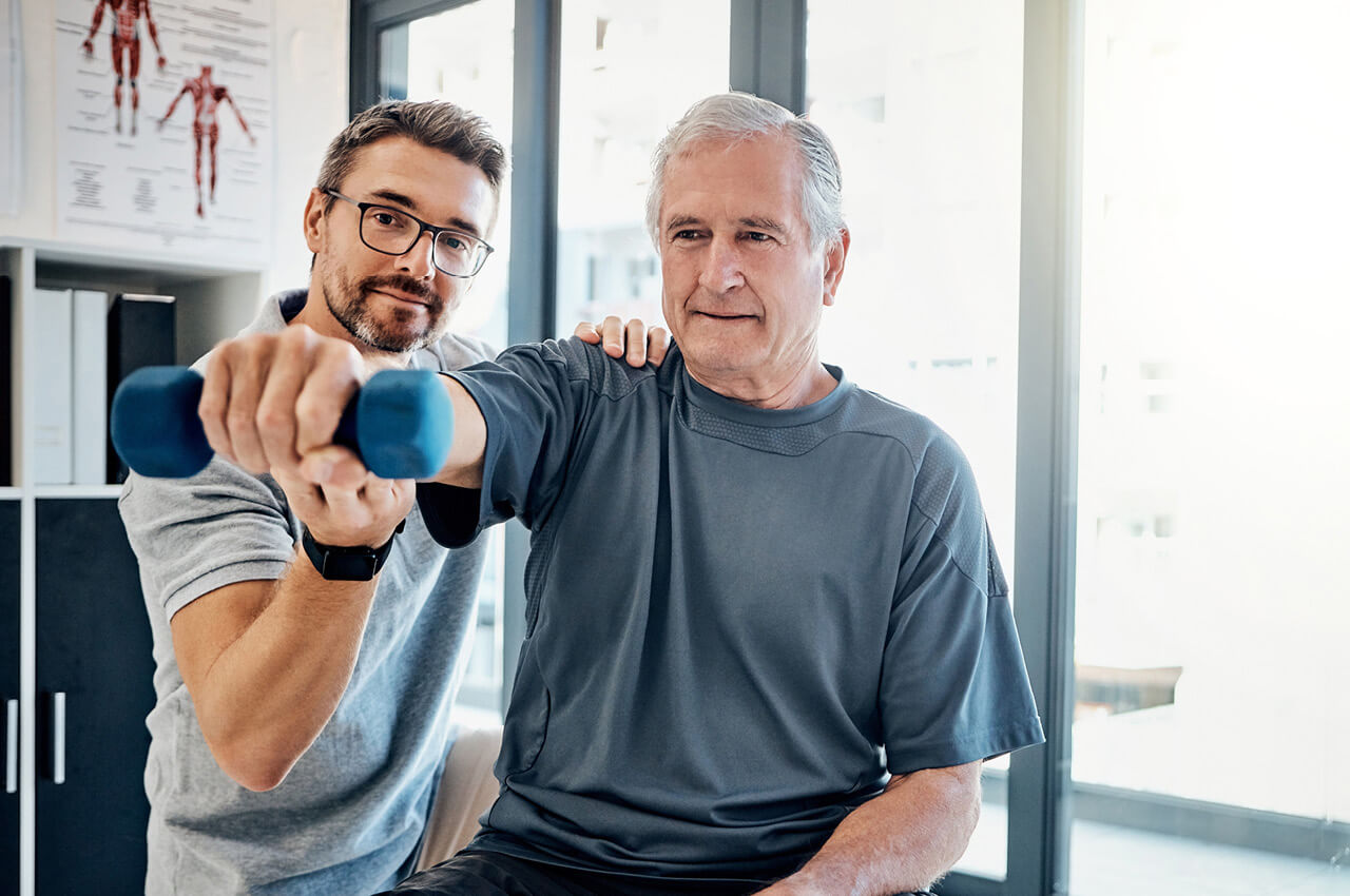 Ein Physiotherapeut (Mann mit Brille und grauem Polohemd) unterstützt und korrigiert einen älteren, männlichen Patienten in einem Trainingsraum bei einer Schulterübung mit einer blauen Hantel. Der Therapeut legt dem Patienten eine Hand auf die Schulter, während dieser die Hantel mit ausgestrecktem Arm hält. Im Hintergrund sind Akten und ein helles Fenster zu sehen.