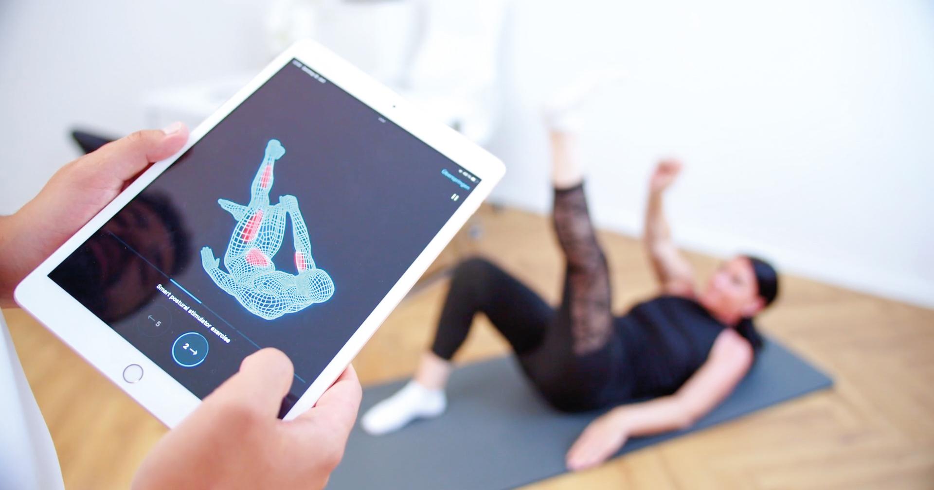 Eine Frau liegt auf einer Sportmatte und übt eine Bewegung aus während eine Hand ein Tablet hält und sich die Übung darauf ansieht