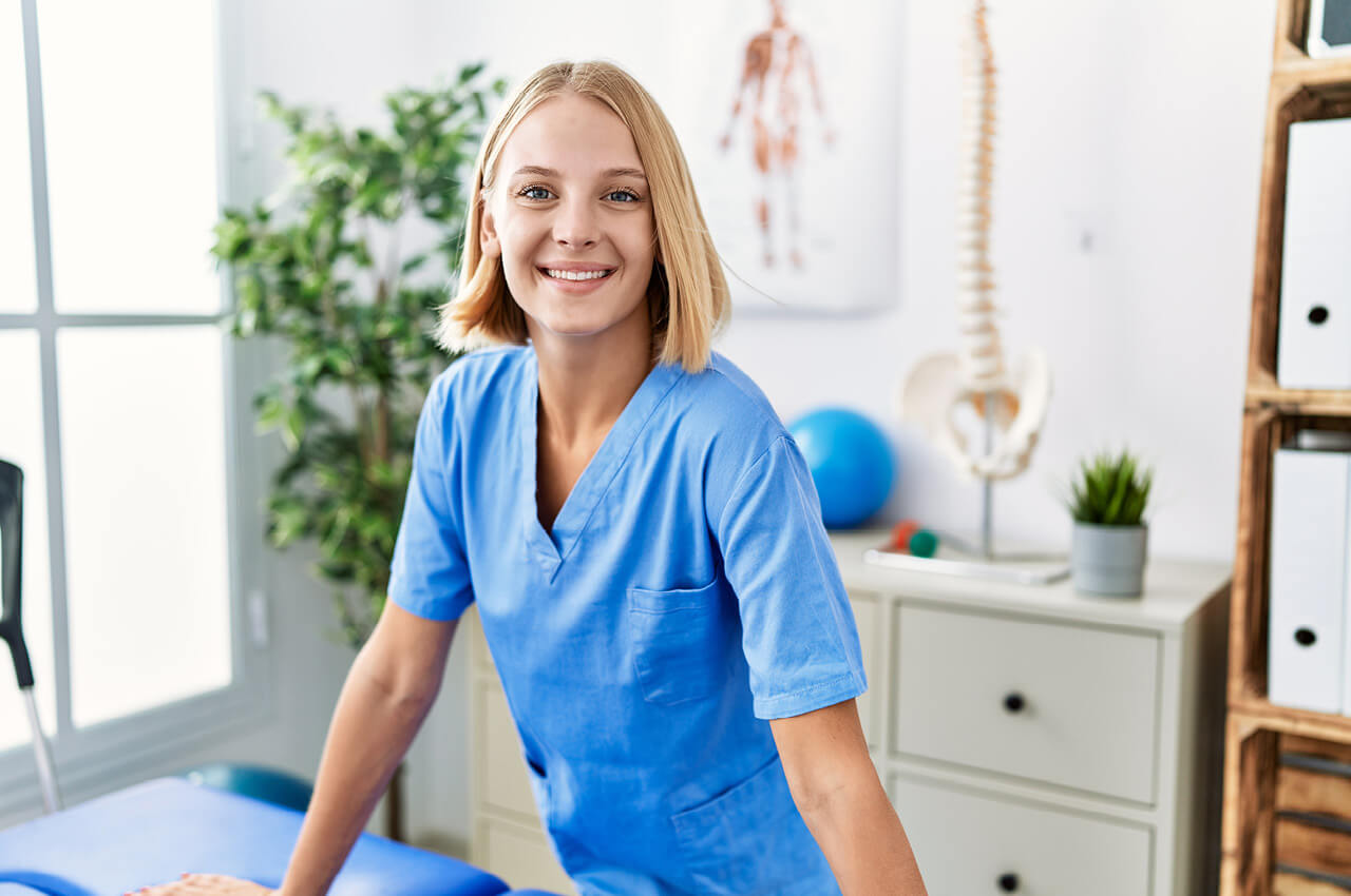 New Pay – Neue Gehaltsstrategie in der therapeutischen Praxis - 2 Eine lächelnde, junge Physiotherapeutin mit kurzem, blondem Haar und blauer Berufskleidung (Kasack) blickt in die Kamera. Sie steht in einem hellen Behandlungsraum, gestützt an einer blauen Therapieliege. Im Hintergrund sind ein Fenster, eine Zimmerpflanze, ein Wirbelsäulen-Skelettmodell und ein anatomisches Poster zu sehen.