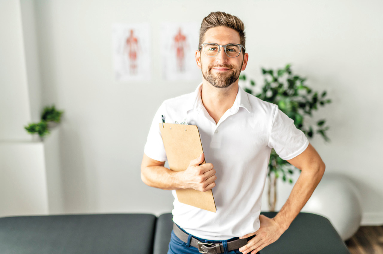 Ein Physiotherapeut hat ein Klemmbrett in der Hand und blickt lächeln in die Kamera. 