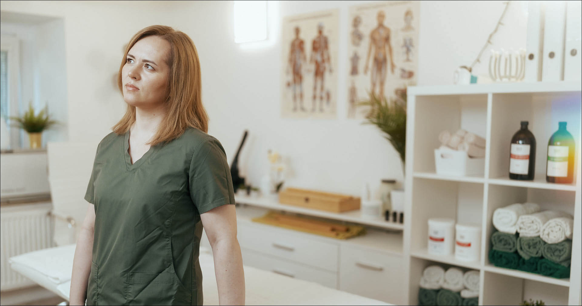 Eine Physiotherapeutin in dunkelgrüner Arbeitskleidung steht mit nachdenklichem, leicht besorgtem Blick in einer Physiotherapiepraxis und schaut zur Seite. Im hellen, leicht unscharfen Hintergrund ist die Praxiseinrichtung zu sehen, darunter eine Behandlungsliege, anatomische Poster an der Wand sowie ein weißes Regal mit Handtüchern und Flaschen.