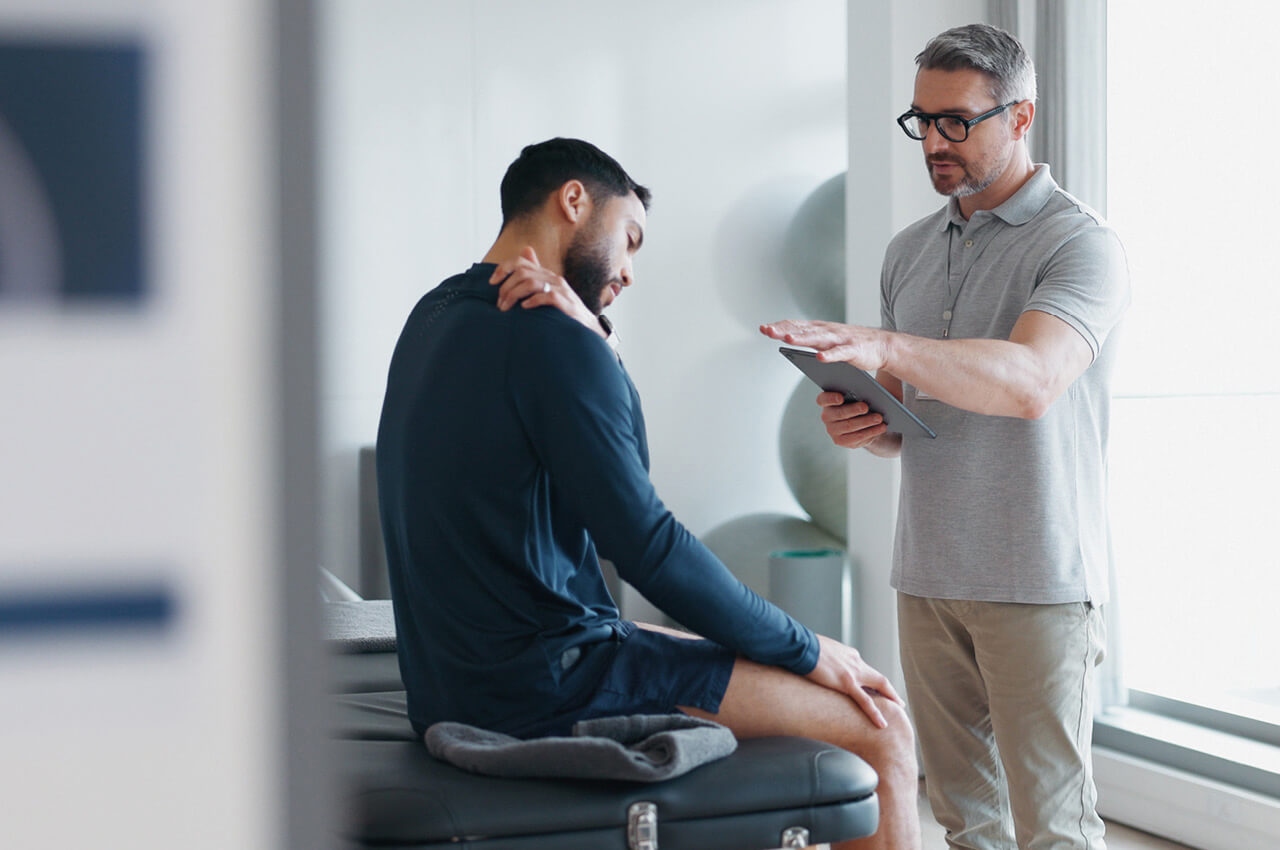 Physiotherapeut nutzt ein Tablet, um einem Patienten mit Schulterschmerzen Befunde oder &Uuml;bungen zu erkl&auml;ren.