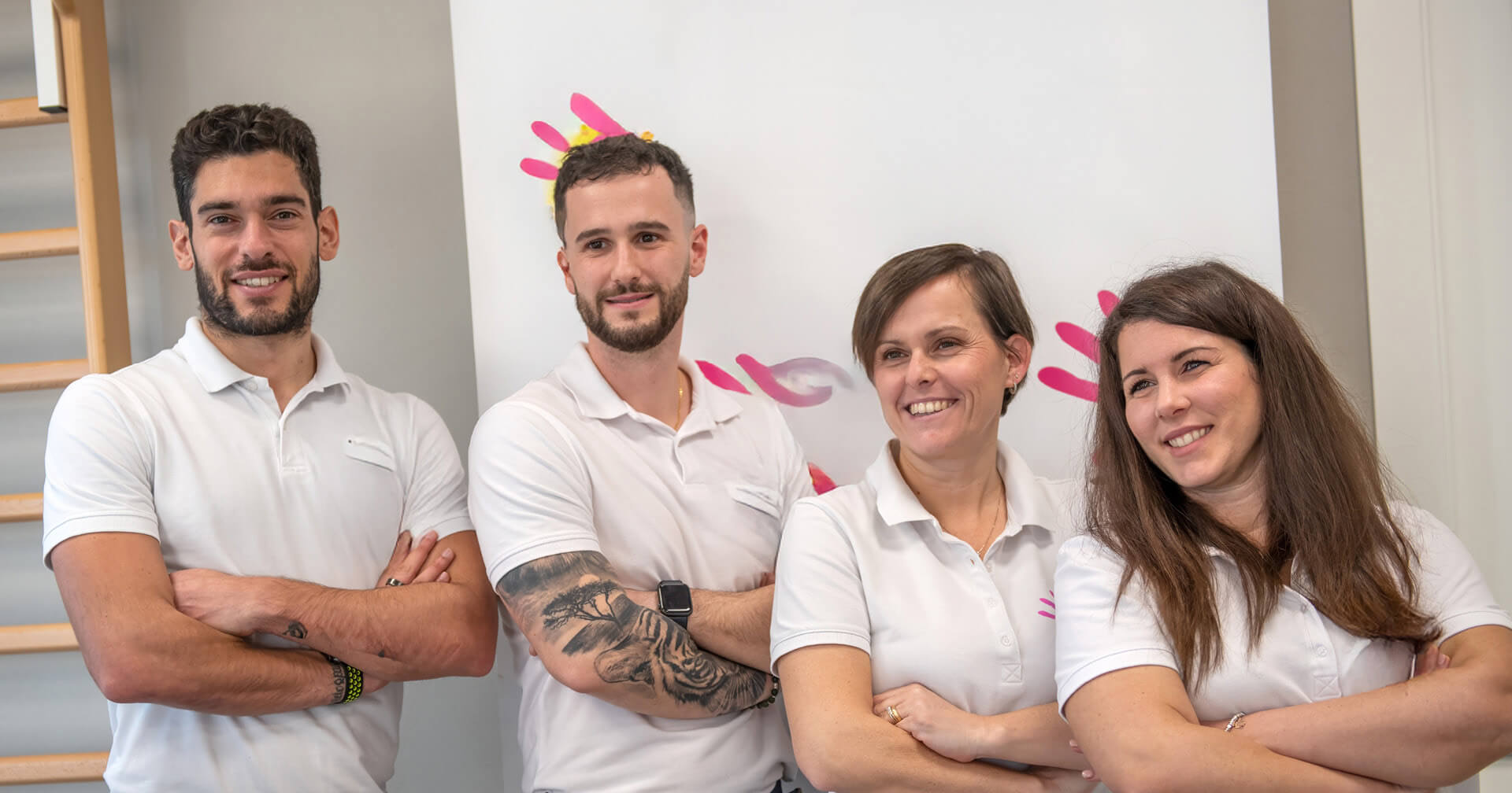 Ein Gruppenporträt von vier lächelnden Teammitgliedern einer Physiotherapiepraxis – zwei Männer und zwei Frauen –, die nebeneinander stehen und die Arme vor der Brust verschränkt haben. Alle tragen weiße Polo-Shirts als Praxisbekleidung.