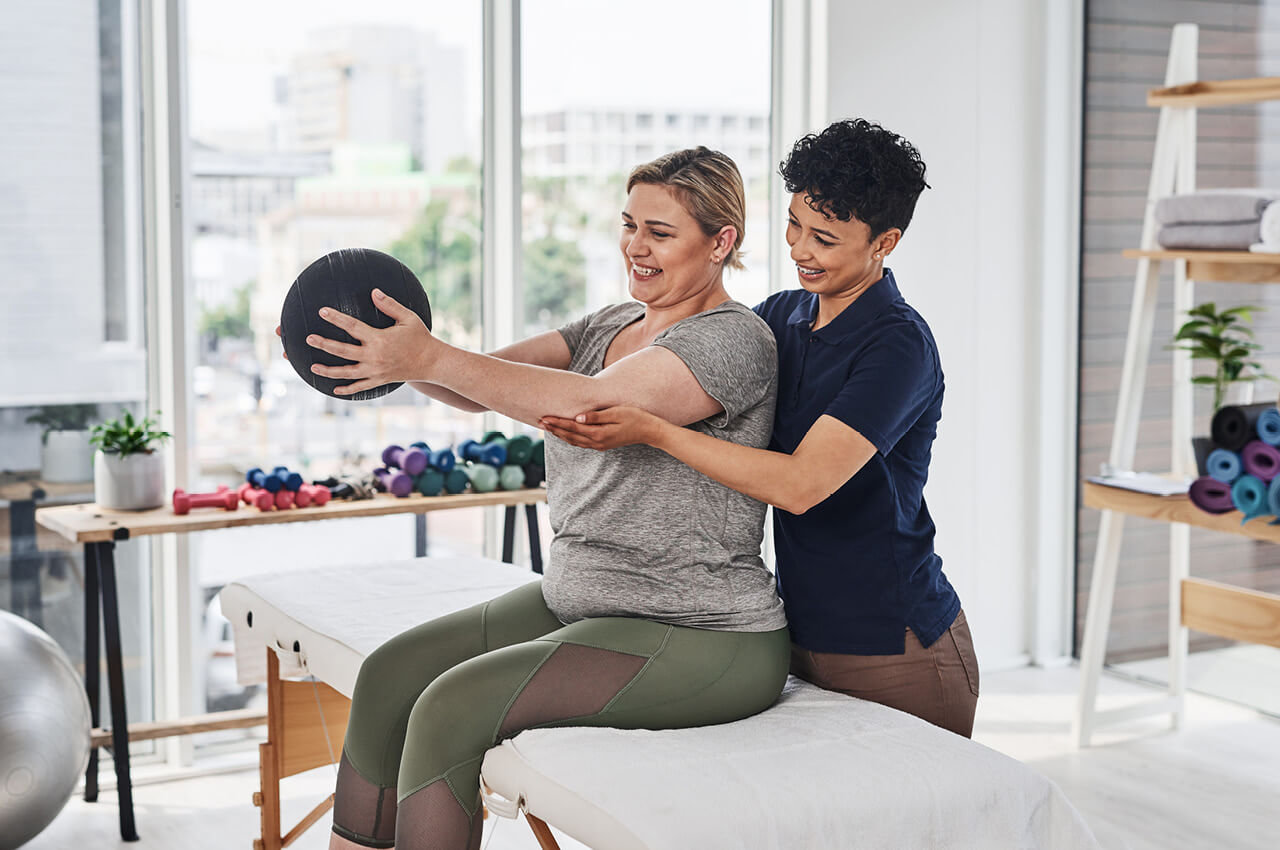 Eine lächelnde Physiotherapeutin (dunkle, kurze Locken, dunkles Poloshirt) unterstützt und leitet eine Patientin (helle, mittellange Haare, graues T-Shirt, grüne Leggings) bei einer Kraftübung mit einem Medizinball. Die Patientin sitzt auf einer Behandlungsliege und hält den schwarzen Medizinball vor sich, während die Therapeutin sie von hinten an den Armen festhält und stützt. Im hellen Hintergrund ist ein großes Fenster mit Blick auf eine Stadt zu sehen.