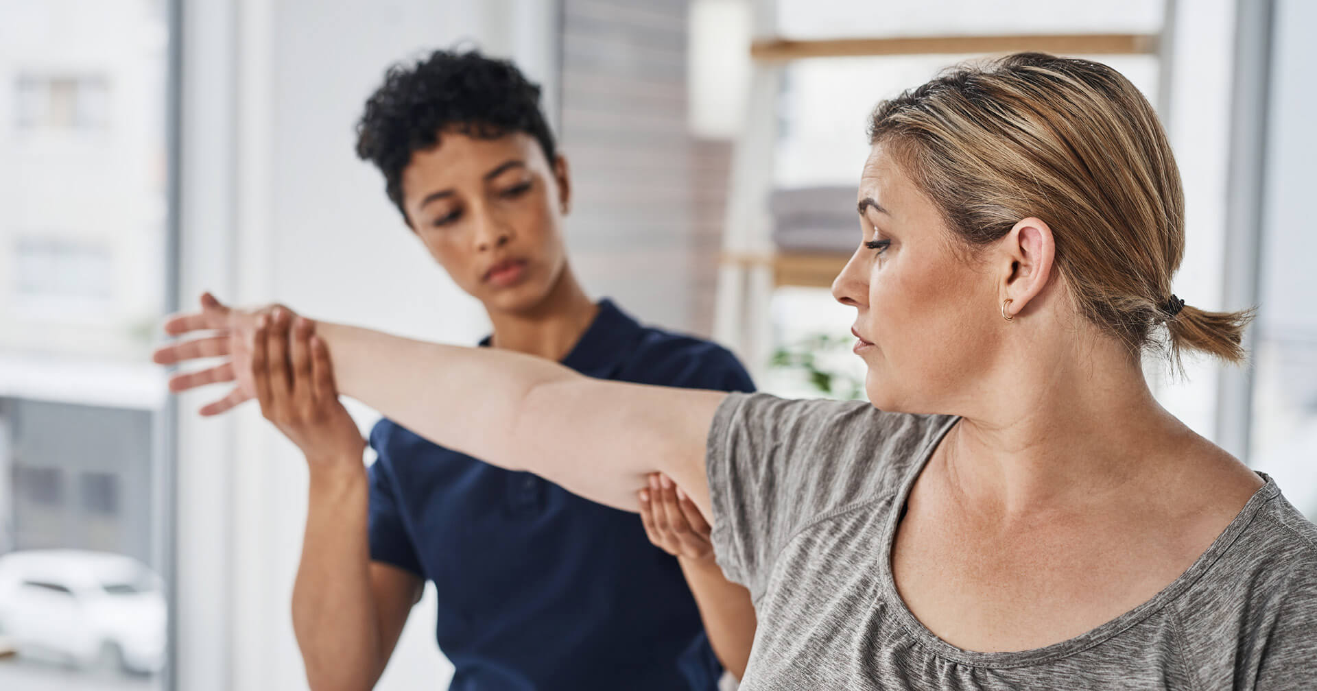 Eine Therapeutin (dunkle, kurze Haare, dunkelblauer Kasack, unscharf im Hintergrund) führt eine Beweglichkeitsübung mit dem Arm einer Patientin (helle, mittellange Haare, graues T-Shirt) durch. Die Patientin blickt mit konzentriertem Gesichtsausdruck zur Seite, während die Therapeutin ihren Arm hält, um ihn zu dehnen oder zu mobilisieren. Die Szene spielt in einem hellen Behandlungsraum mit großen Fenstern.