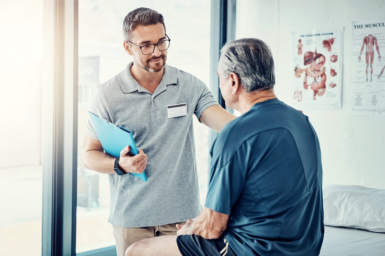 Ein lächelnder, junger Physiotherapeut oder Arzt mit Brille und grauem Polohemd, hält ein Klemmbrett und legt dem älteren, männlichen Patienten in einem Behandlungsraum tröstend die Hand auf die Schulter. Der Patient sitzt auf einer Behandlungsliege und hat den Rücken zum Betrachter gewandt. Im Hintergrund sind ein großes Fenster und medizinische Plakate mit anatomischen Darstellungen zu sehen.